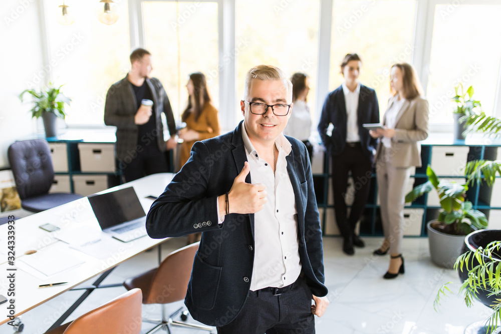 Team in the office. Young handsome businessman standing in the foreground showing thumb up, his team of co-workers in the background Team in the office. Young handsome businessman standing in the foreground showing thumb up, his team of co-workers in the background