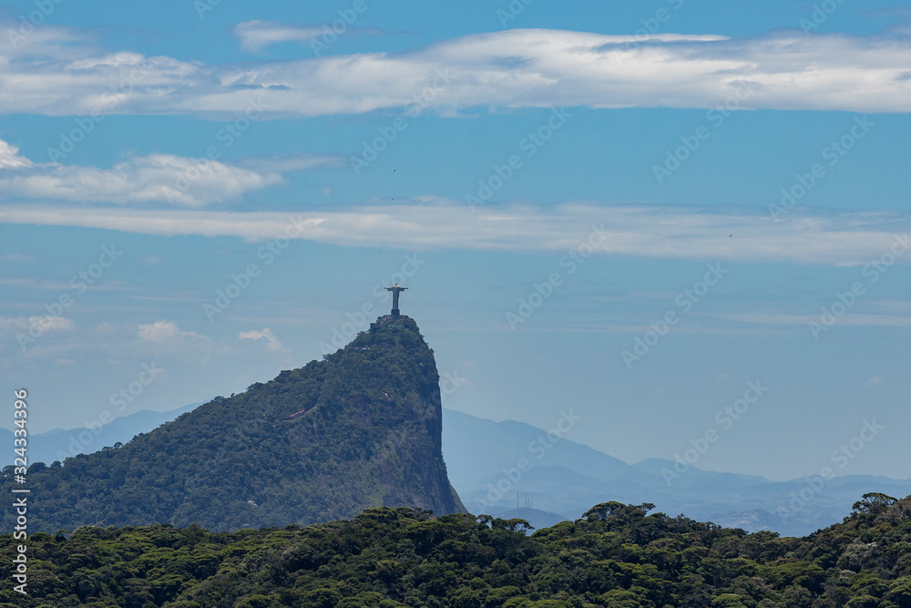 Corcovado mountain in Rio de Janeiro, Brazil, seen from the Pedra Bonita viewpoint with part of the Tijuca forest in the foreground and mountain peak and Christ statue in the back against a blue sky