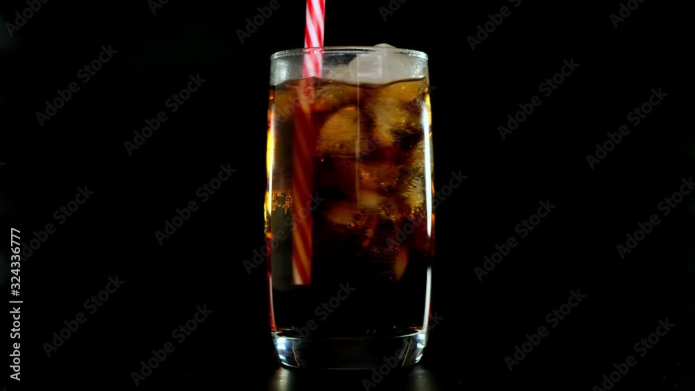 Putred straw into cola with ice cubes in glass over black background. Close up.