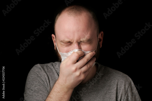 Fototapeta Naklejka Na Ścianę i Meble -  Young guy with a beard In a medical mask sneezes, covering her mouth with her hands ob black background. Seasonal infection, colds and runny nose, coronavirus.