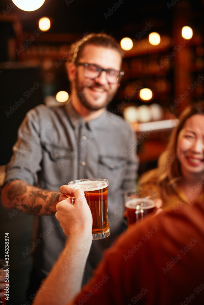 Smiling hipster man standing at pub with friends and passing pint of beer to his friend. Group of friends having fun at pub. Nightlife.