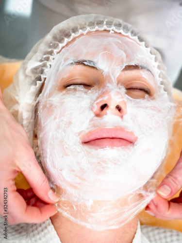 Girl beautician applies a to rejuvenate a mask on the face of an attractive girl in a spa salon. Beautiful woman during rejuvenation procedure. Face treatment at cosmetic clinic.