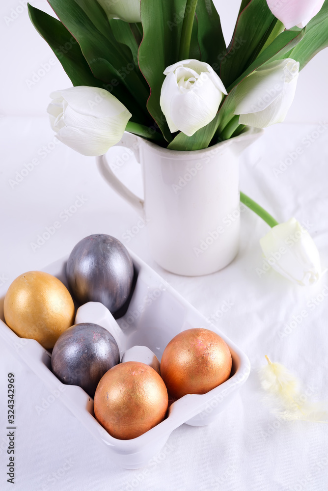 Golden Easter Eggs in ceramic egg tray with tulip on white table. Easter background or easter concept. Golden Easter Eggs in ceramic egg tray with tulip on white table. Easter background or easter concept.