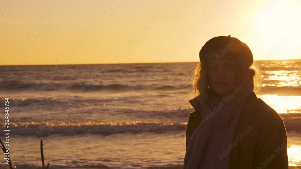 Slow motion of a young brunette model woman with a fringe and a black vintage basque cap at a beach in Florida by the golden light of the evening sun by sunset with flares making eye contact and waves Slow motion of a young brunette model woman with a fringe and a black vintage basque cap at a beach in Florida by the golden light of the evening sun by sunset with flares making eye contact and waves