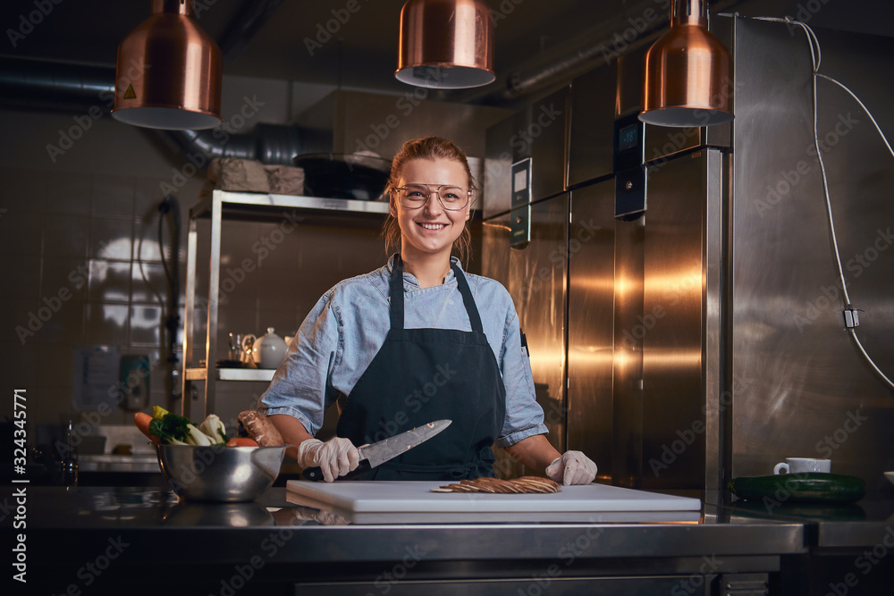 Joyful and excited female chef standing in a dark cafe kitchen next to big bowl of fresh vegetables and cutting board with mushroom pieces on it, holding a knife, wearing apron and denim shirt, posing Joyful and excited female chef standing in a dark cafe kitchen next to big bowl of fresh vegetables and cutting board with mushroom pieces on it, holding a knife, wearing apron and denim shirt, posing