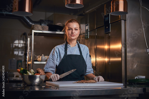 Confident and serious woman chef standing in a dark kitchen next to a big bowl of fresh vegetables and cutting board with mushroom pieces on it  holding a kitchen knife  wearing apron and denim shirt