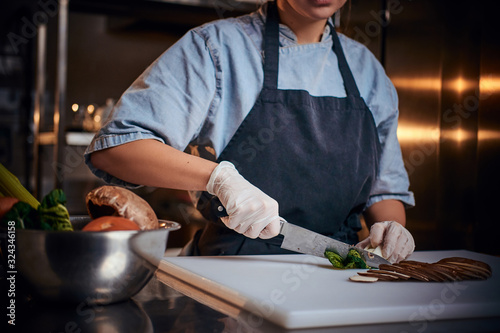 Chef female standing on a dark restaurant kitchen  wearing gloves  apron and denim shirt  cutting vegetables on a cutting board net to the bowl with raw vegetables  close up view