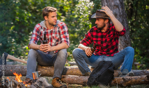 nice speak. man drink beer at bonfire. ranger at outdoor activities. spend picnic weekend in nature. Adventure concept. hike and people. two men relax at fire. hiking and camping. male friendship