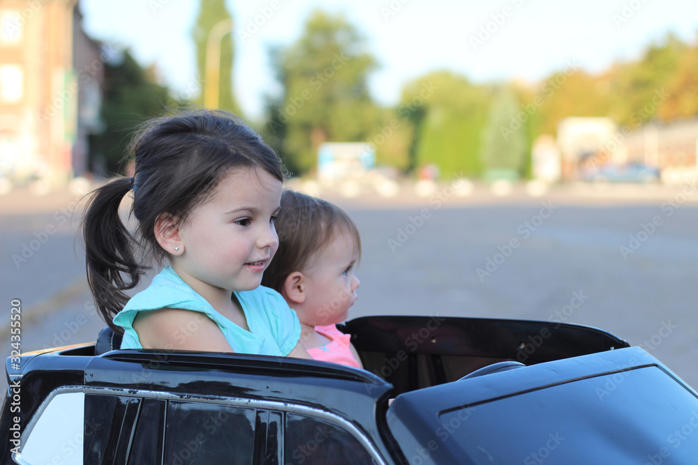 Two amazing girls ride in one big toy car on city street asphalt. Outdoor driving in a summer attraction for children Two amazing girls ride in one big toy car on city street asphalt. Outdoor driving in a summer attraction for children