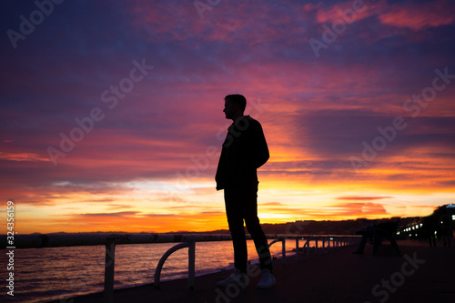 Guy looking at gorgeous sunset on the shore of sea in Nice, France. Black silhouette of man on gorgeous sunset with orange, purple and rose clouds. 