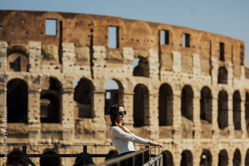 Happy girl in white shirt with glasses smiling and admires of Rome Colosseum. She standing near Coliseum, Rome, Italy. The girl thinking about history at the Coliseum.