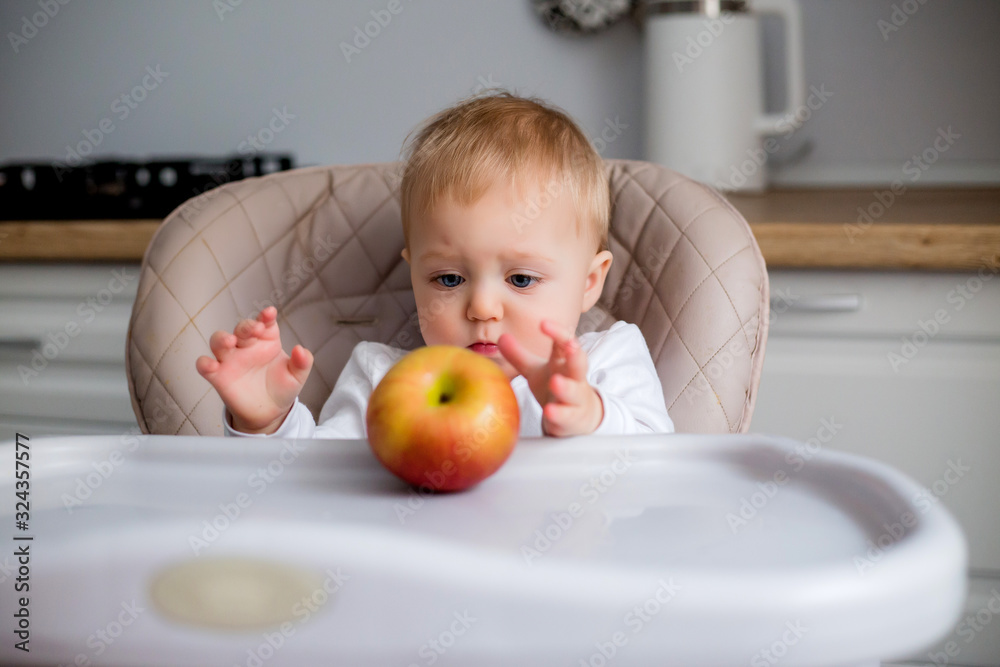 baby boy 7 months old sitting in a high chair in the kitchen and eating an Apple baby boy 7 months old sitting in a high chair in the kitchen and eating an Apple