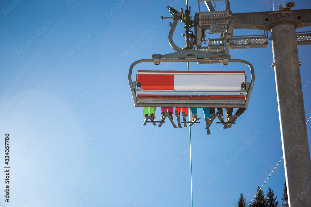 The selective focus of the skier's legs in the ski lift, the sun illuminates the six-seat ski lift. Below the cable car there is a ski slope and skis and a pine forest around. The selective focus of the skier's legs in the ski lift, the sun illuminates the six-seat ski lift. Below the cable car there is a ski slope and skis and a pine forest around.
