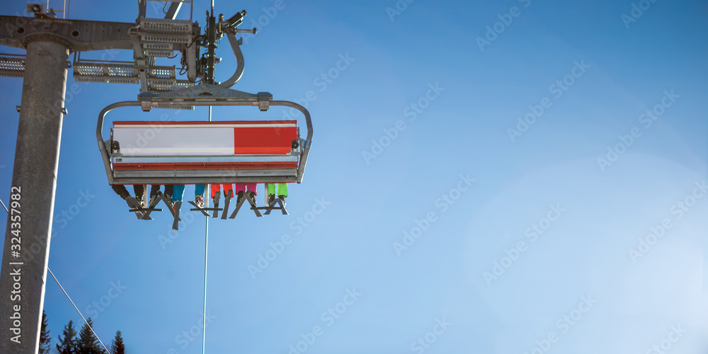 The selective focus of the skier's legs in the ski lift, the sun illuminates the six-seat ski lift. Below the cable car there is a ski slope and skis and a pine forest around. The selective focus of the skier's legs in the ski lift, the sun illuminates the six-seat ski lift. Below the cable car there is a ski slope and skis and a pine forest around.