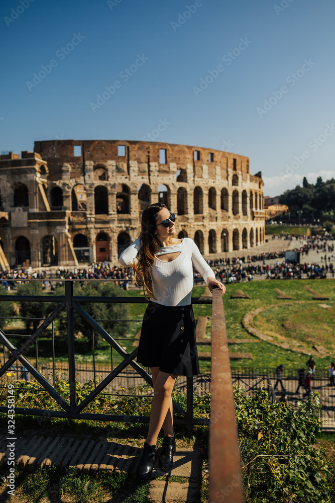 Brunette girl with long hair in dress sits on the background of the majestic ancient Colosseum in Rome. Great Colosseum. Rome, Italy. Traveler girl looking on Coliseum. 