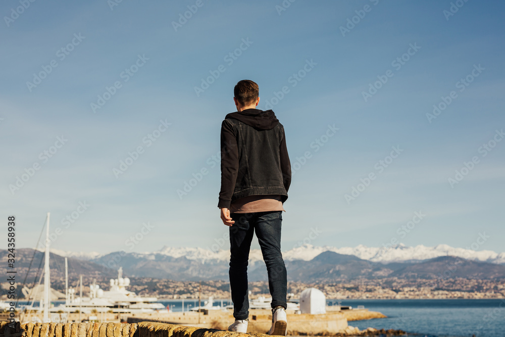Back view of man. He walk on the waterfront and enjoying the view of the sea and landscape snowing mountain in Antibes, France. Port city on the background.