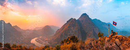 Sunset panoramic view of couple of trekkers sitting on a rock on top of Nong Khiaw View Point with beautiful mountain and Nam Ou river in background, Luang Prabang Province, Northern Laos. photo