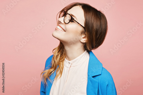 Happy young woman in stylish glasses and trendy blue coat, isolated at pink. Cheerful model girl in pink blouse and blue coat, and modish eyewear over pink background, isolated. Copy space. Fashion