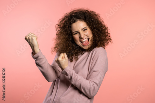 Girl with curly hair and extraordinary makeup shows yes gesture of victory, she achieved result, goals. Surprised excited happy woman on pink background