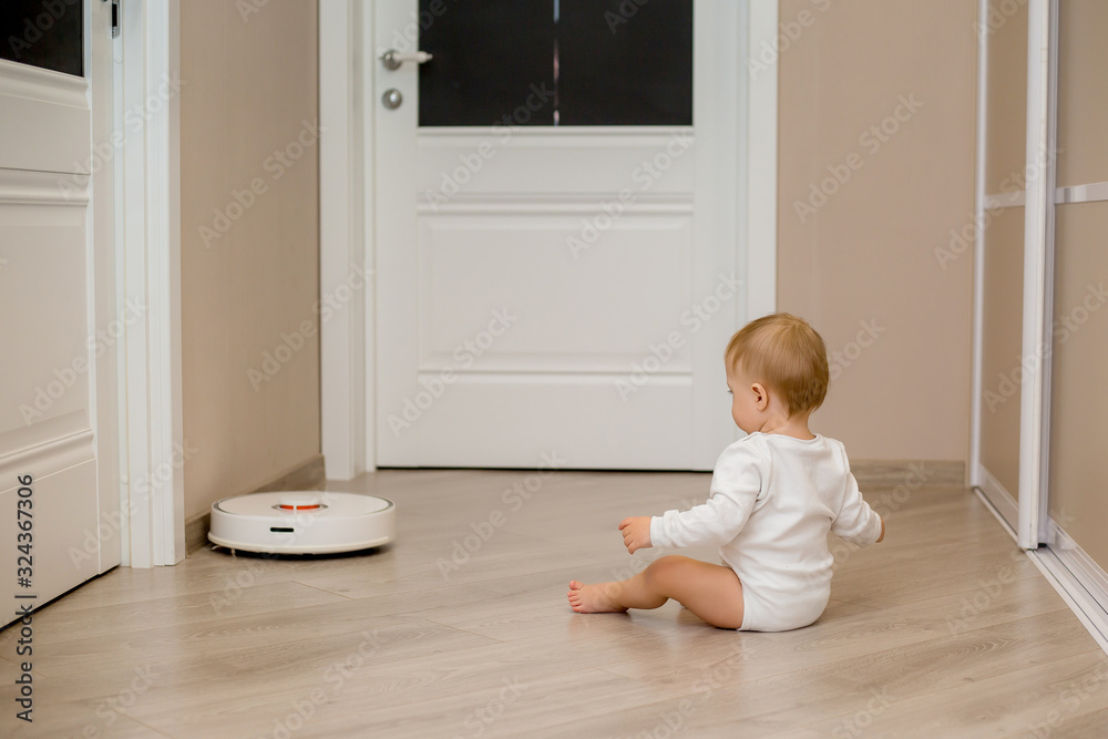 baby boy in white clothes with a robotic vacuum cleaner on the floor of the house baby boy in white clothes with a robotic vacuum cleaner on the floor of the house