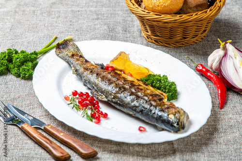 Trout with lemon and herbs. On the plate is a trout with a head, grilled. Near lemon, parsley and red currant. Plate on a gray canvas. Next to the red pepper, fork, spoon, bread basket photo