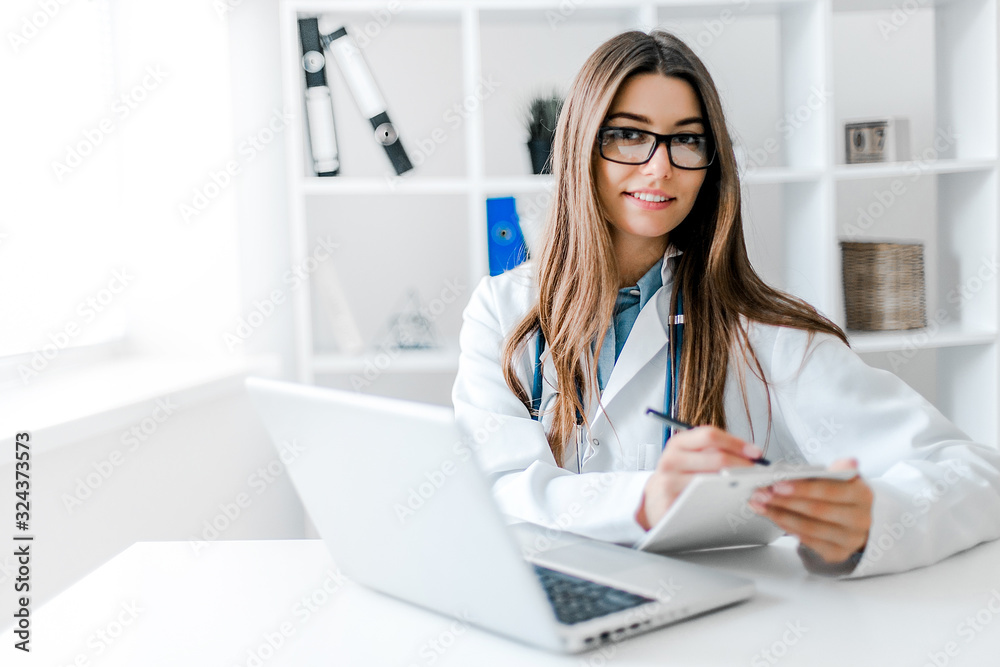 Smiling young female doctor working at the clinic , she is using a computer and writing medical reports. Smiling young female doctor working at the clinic , she is using a computer and writing medical reports.