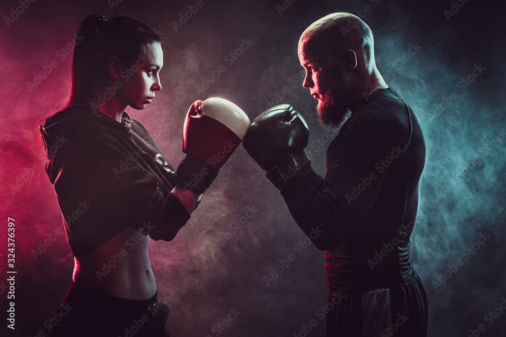 Fototapeta Woman exercising with trainer at boxing and self defense lesson, studio, smoke on background. Aggresively look each other. Stand in front.