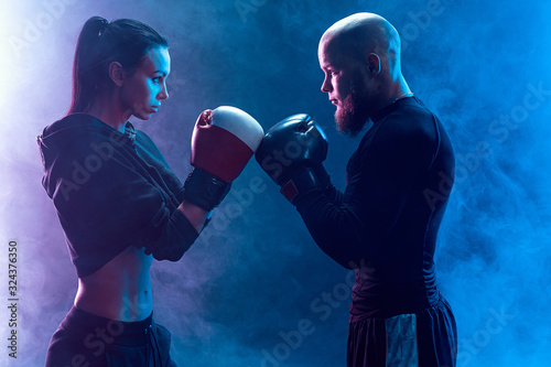 Woman exercising with trainer at boxing and self defense lesson, studio, smoke on background. Aggresively look each other. Stand in front. © zamuruev