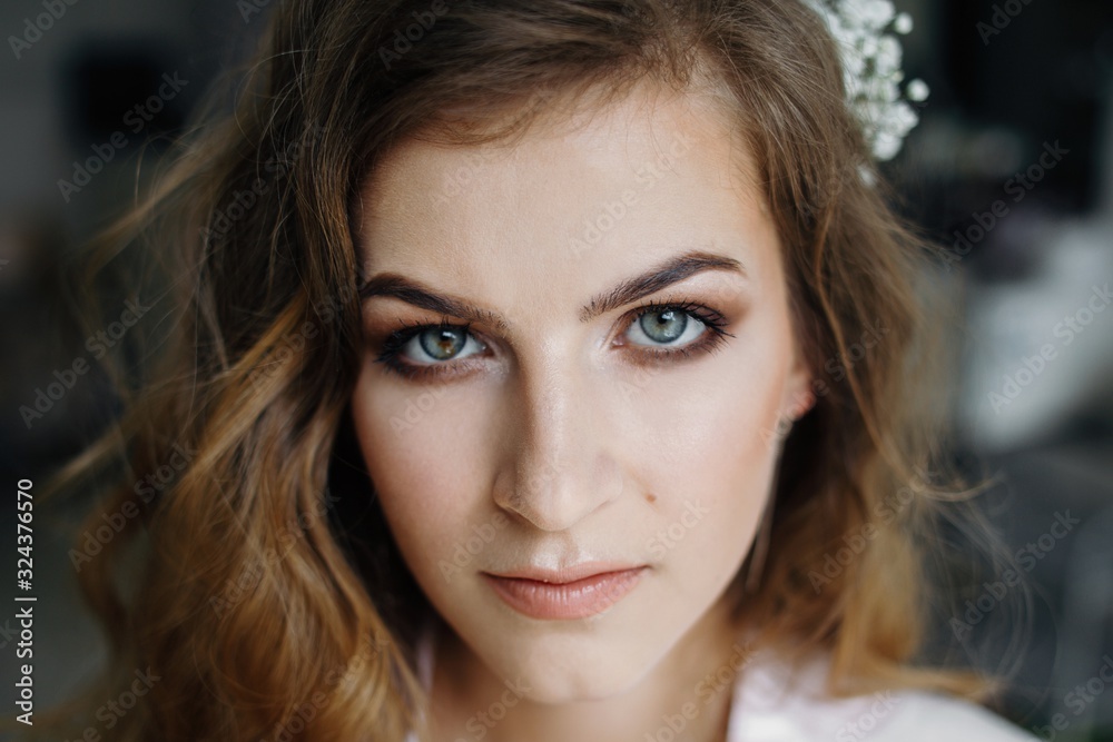 Naklejka premium Close up of attractive woman with beautiful blue eyes and nature make up, curly hairstyle and flower in hair, seriously looking at camera.