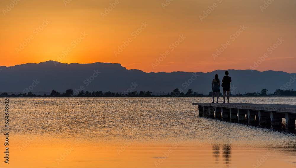 A young couple watches the sunset on a pier at the mouth of the Ebro river, Trabucador beach, in the Mediterranean Sea, in the province of Tarragona (Spain).