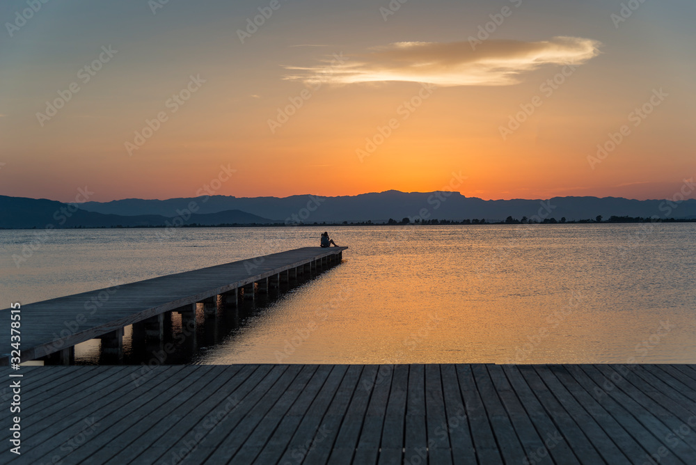 Fototapeta premium A young couple watches the sunset on a pier at the mouth of the Ebro river, Trabucador beach, in the Mediterranean Sea, in the province of Tarragona (Spain).