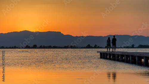 Fototapeta Naklejka Na Ścianę i Meble -  A young couple watches the sunset on a pier at the mouth of the Ebro river, Trabucador beach, in the Mediterranean Sea, in the province of Tarragona (Spain).