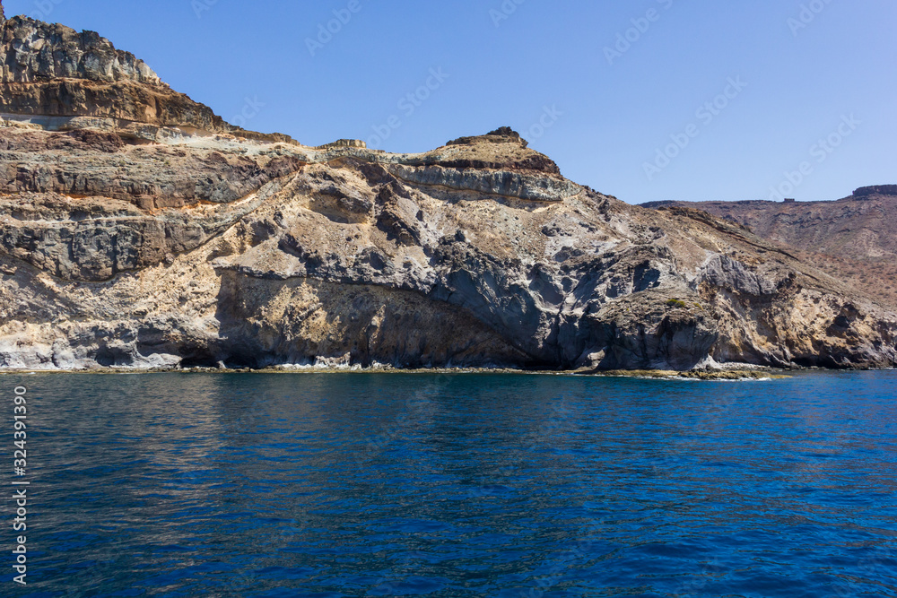 Fototapeta premium Arid rocky landscape by ocean in Gran Canaria, Spain. Canary Islands volcanic coast view from sea. Boat trip, summer vacation concepts