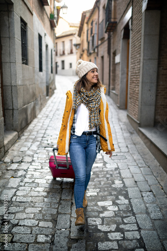young girl sightseeing through the streets of a city with yellow coat, wool cap and suitcase © Sergio