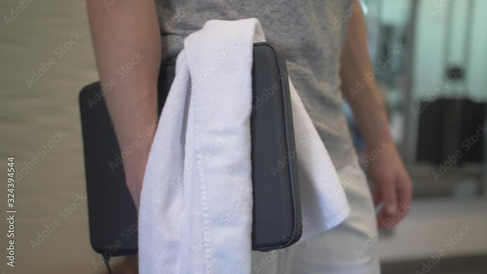 A close up of a man walking in the gym with fitness clothing, holding a black laptop case and a white towel. Fitness while traveling and on business trip. Slow motion. A close up of a man walking in the gym with fitness clothing, holding a black laptop case and a white towel. Fitness while traveling and on business trip. Slow motion.