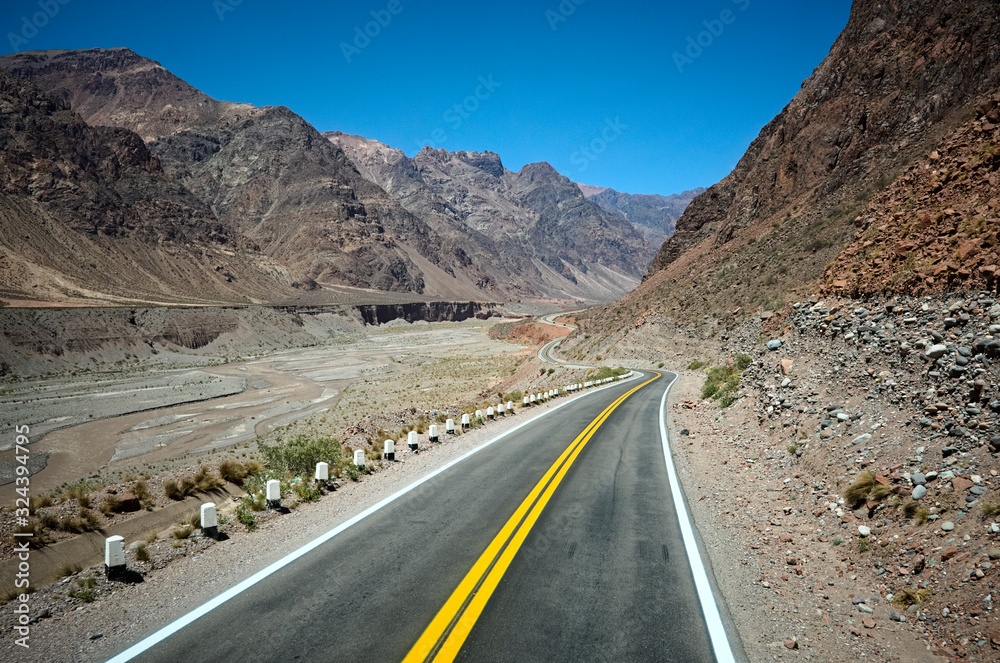 Highway road in the Andes mountains along canyon river. Empty road in arid climate as desert with rocky terrain. Curved road with yellow dividing line through mountains in Mendoza province, Argentina Highway road in the Andes mountains along canyon river. Empty road in arid climate as desert with rocky terrain. Curved road with yellow dividing line through mountains in Mendoza province, Argentina