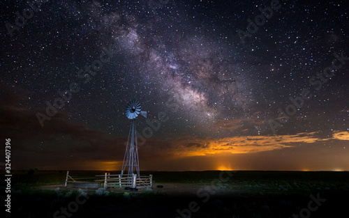 A old fashion windmill on a farm under the night sky. The Milky Way and the stars are visible overhead.