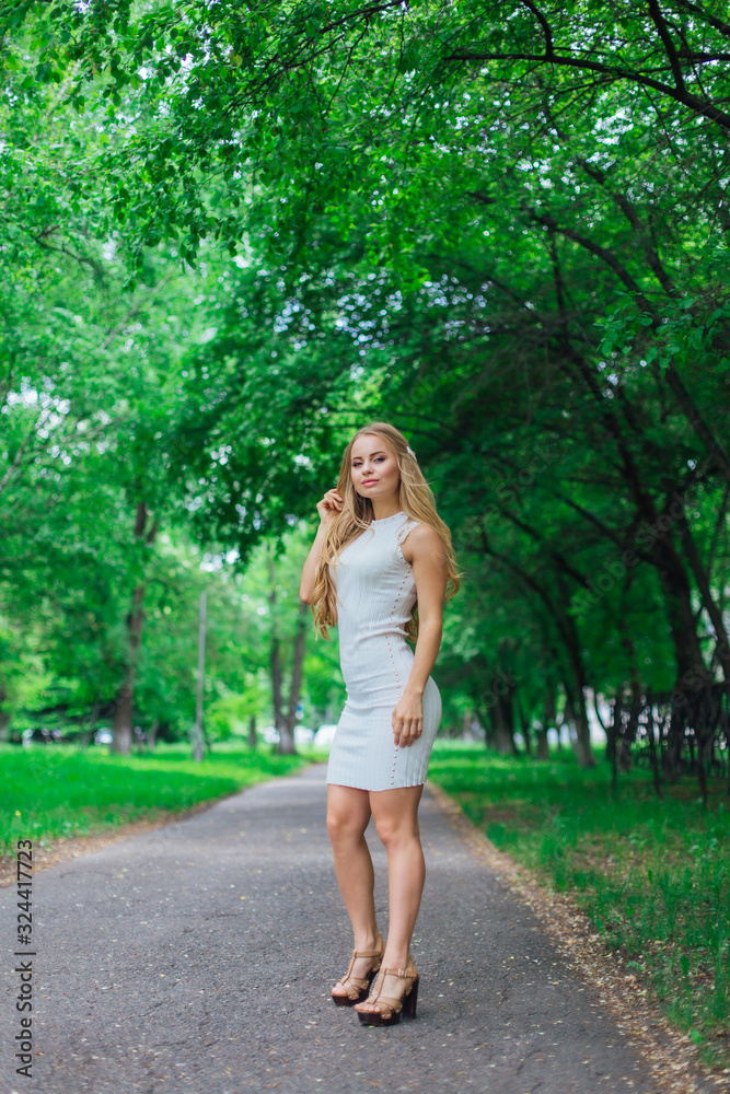 Portrait of a charming blond woman wearing beautiful white dress standing on the road under trees. Portrait of a charming blond woman wearing beautiful white dress standing on the road under trees.