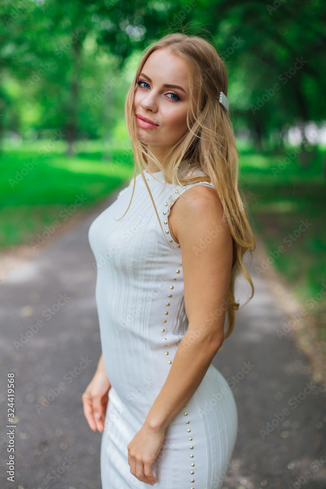 Portrait of a charming blond woman wearing beautiful white dress standing on the road under trees. Portrait of a charming blond woman wearing beautiful white dress standing on the road under trees.