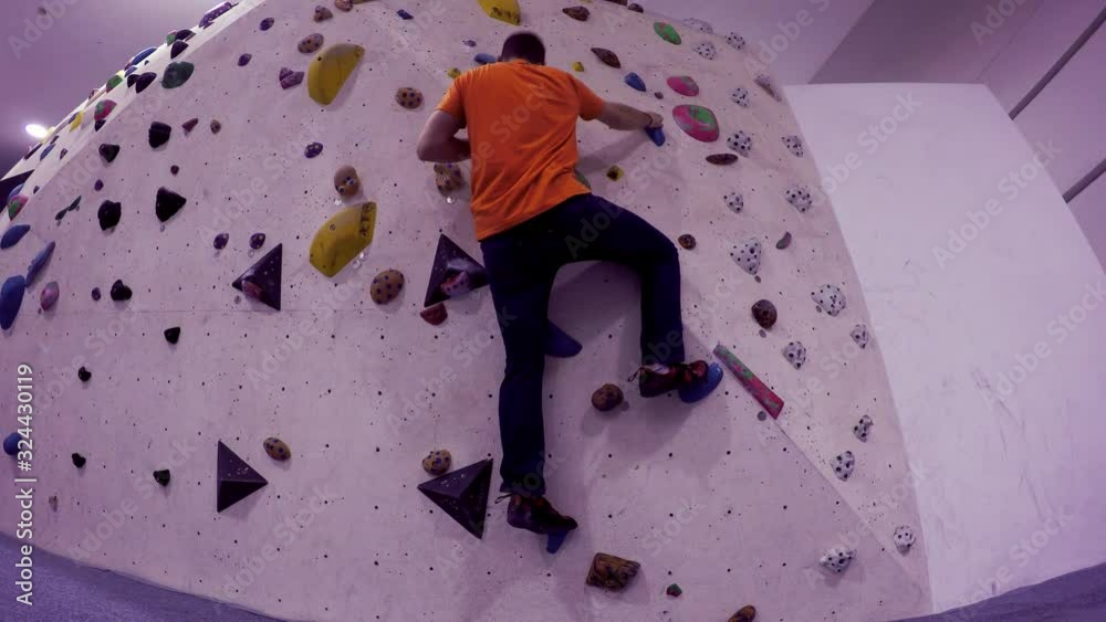 Boulder gym climbing wall seen from below as a climber approaches and climbs up and then down with no rope. The wall has a pointing angle sticking out. The route taken up is the one with blue handles. Boulder gym climbing wall seen from below as a climber approaches and climbs up and then down with no rope. The wall has a pointing angle sticking out. The route taken up is the one with blue handles.