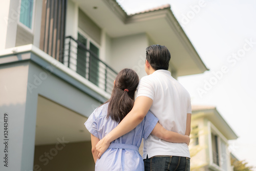 Back portrait of Asian young couple standing and hugging together looking happy in front of their new house to start new life. Family, age, home, real estate and people concept..