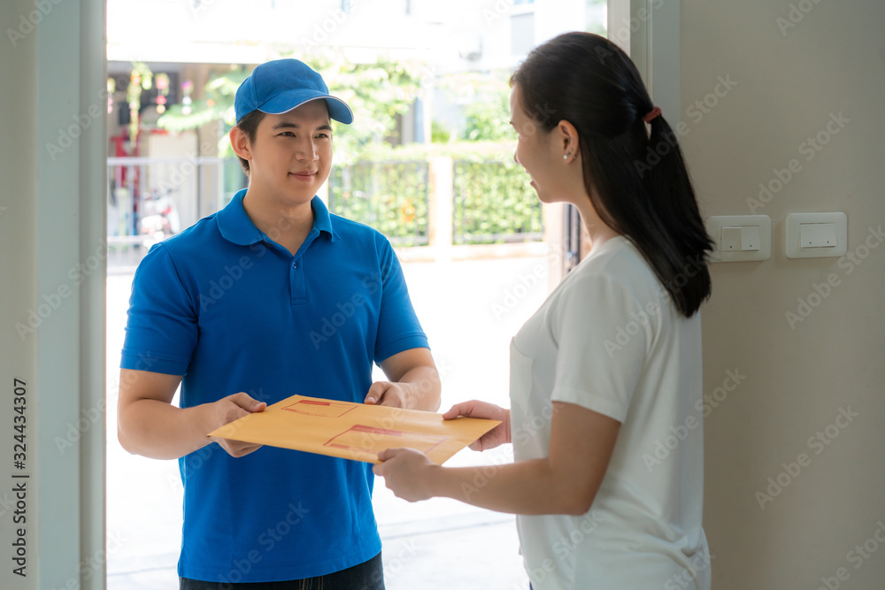 Asian delivery young man in blue uniform smile and holding letter or document envelope in front house and Asian woman accepting a delivery of envelope from deliveryman. Advertising, Transportation. Asian delivery young man in blue uniform smile and holding letter or document envelope in front house and Asian woman accepting a delivery of envelope from deliveryman. Advertising, Transportation.