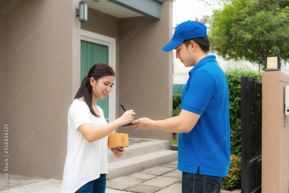 Asian delivery young man in blue uniform smile and holding smartphone in front house and Asian woman sign signature in smartphone to accepting a delivery of boxes from deliveryman.. Asian delivery young man in blue uniform smile and holding smartphone in front house and Asian woman sign signature in smartphone to accepting a delivery of boxes from deliveryman..