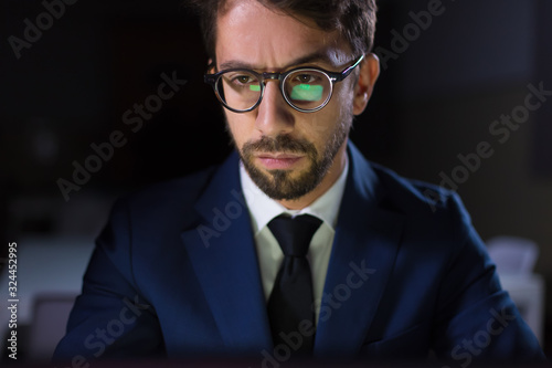 Handsome young businessman working with laptop at night. Front view of bearded man with reflection of screen on glasses. Overwork, working late concept