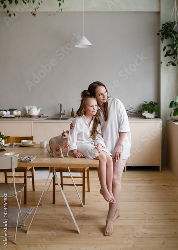 Happy mother and daughter in home white shirts relax in a hammock, have Breakfast and drink tea in the kitchen, have fun, communicate, hug in a cozy bright house, comfort, love, care, family, nature
