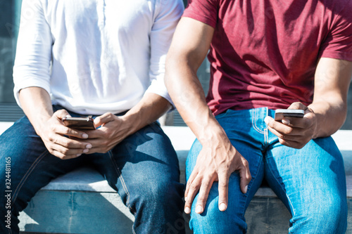 Closeup of two men in casual using smartphones outside. Cropped portrait of two male friends in casual sitting on parapet outdoors and holding mobile phones. Communication concept