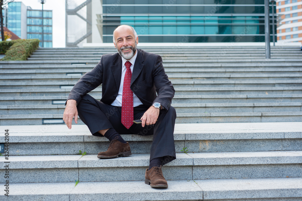 Smiling businessman looking at camera while sitting on stairs. Successful cheerful middle aged attorney holding modern phone. Technology, business concept Smiling businessman looking at camera while sitting on stairs. Successful cheerful middle aged attorney holding modern phone. Technology, business concept