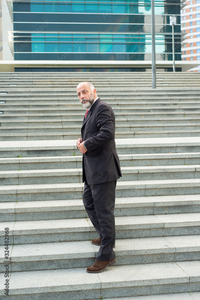 Full length portrait of confident businessman standing on stairs. Successful mature business consultant fastening button of suit. Concept of confidence Full length portrait of confident businessman standing on stairs. Successful mature business consultant fastening button of suit. Concept of confidence