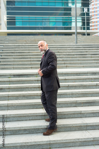 Full length portrait of confident businessman standing on stairs. Successful mature business consultant fastening button of suit. Concept of confidence