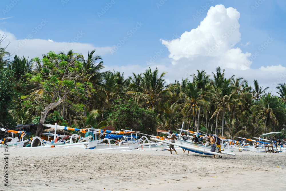 Fishermen carry a boat on a virgin beach in Bali. Fishing boats on white sand. Coconut trees and blue sky with clouds on the background. Clear sunny day
